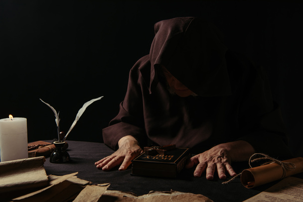 senior abbot in hood sitting near bible and medieval manuscripts isolated on black - Photo, Image