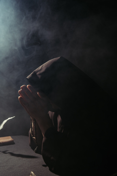 monnik met verduisterd gezicht bidden met rozenkrans 's nachts op zwart met rook - Foto, afbeelding