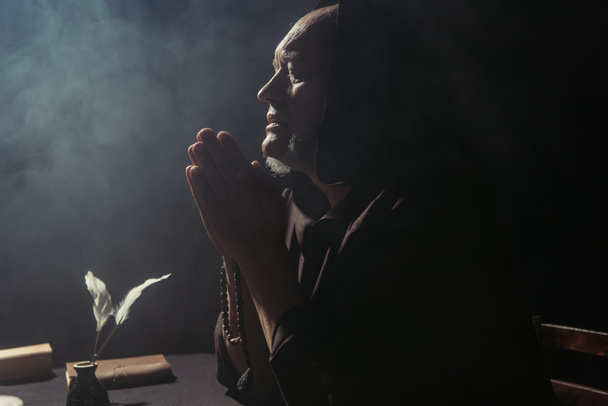monje medieval rezando cerca de tintero y manuscrito sobre fondo negro con humo - Foto, imagen