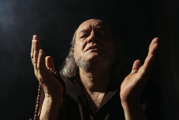 vista frontal do abade sênior com rosário orando com olhos fechados sobre fundo preto - Foto, Imagem