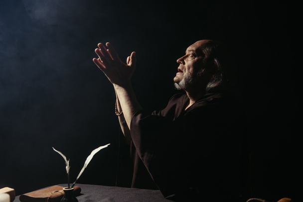 vista lateral del monje medieval rezando cerca de tintero con plumas sobre fondo negro - Foto, imagen