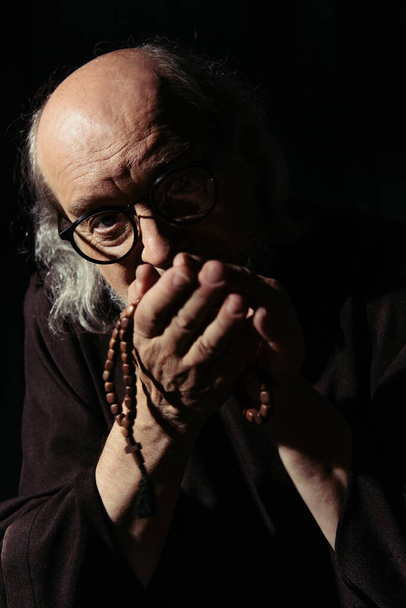 monaco anziano in occhiali tenendo rosario e guardando la fotocamera isolata su nero - Foto, immagini