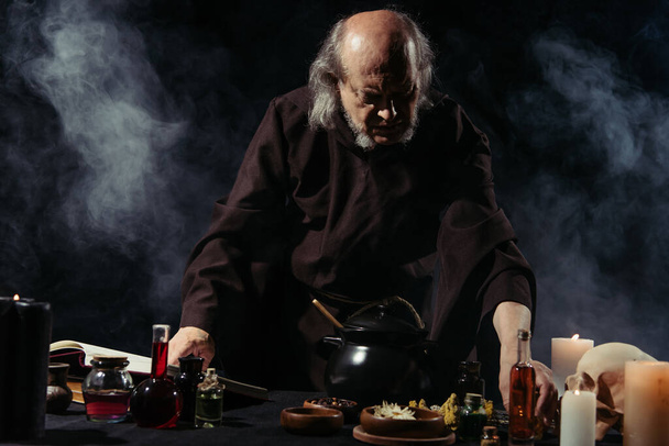 středověký alchymista připravuje lektvar v blízkosti tekutých a sušených složek na černém pozadí s kouřem - Fotografie, Obrázek