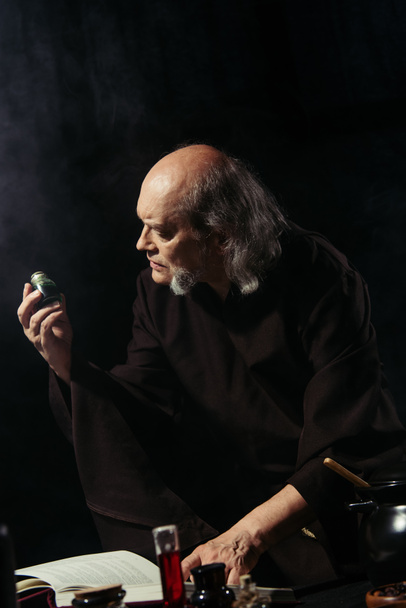 старший алхимик держит флакон с волшебным ингредиентом рядом с колбами и волшебной поваренной книгой, изолированной на черном - Фото, изображение