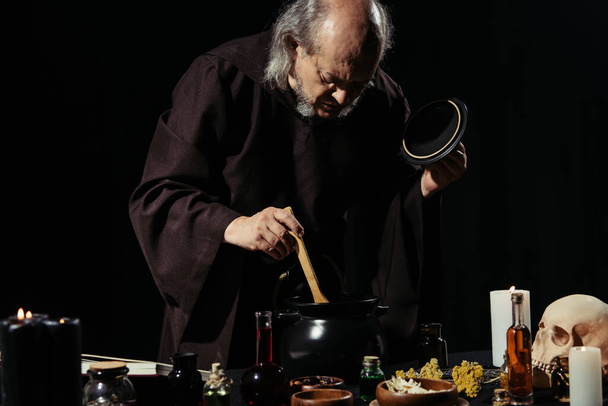 alchemist in black robe preparing potion with herbal and liquid ingredients isolated on black - Photo, Image