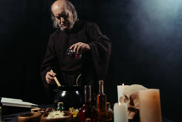 tajemný alchymista přidání složky do hrnce při vaření v tmě na černém pozadí s kouřem - Fotografie, Obrázek