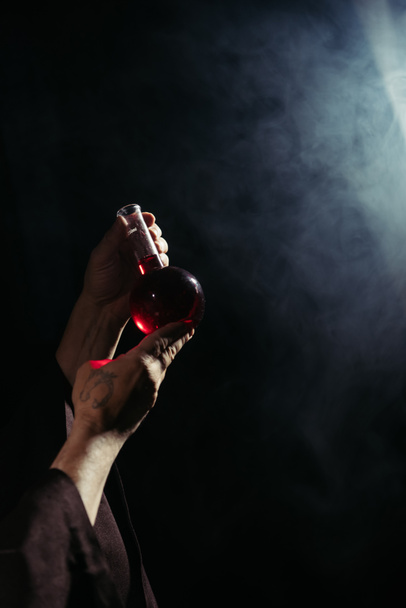 partial view of magician holding flask with potion on black background with smoke - Photo, Image