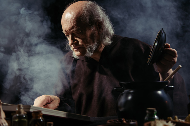 keskiaikainen alkemisti lukee maagista keittokirjaa valmistellessaan taikajuomaa yöllä mustalla taustalla savulla - Valokuva, kuva