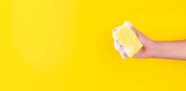 Persons hand holding yellow sponge for dish wash. Washcloth covered in soap. Domestic chores and supplies concept. Sensitive dishwashing detergent. Copy space in left side. Isolated on yellow - Photo, Image