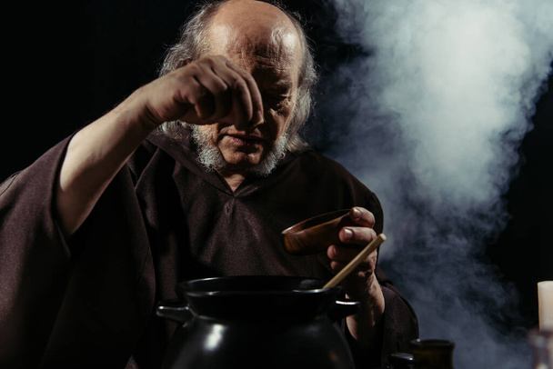 mago anziano aggiungendo ingrediente dalla ciotola in pentola su sfondo nero con fumo - Foto, immagini