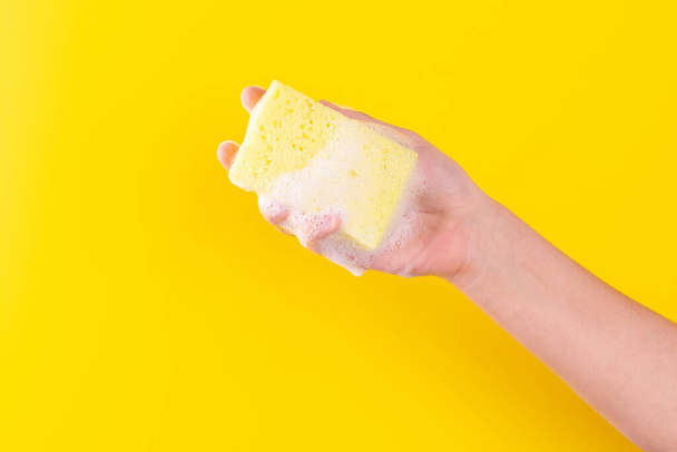 Persons hand holding yellow sponge for dish wash. Washcloth covered in soap. Domestic chores and supplies concept. Sensitive dishwashing detergent. Copy space in left side. Isolated on yellow - Foto, immagini