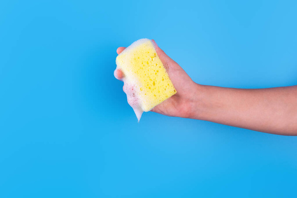 Persons hand holding yellow sponge for dish wash. Washcloth covered in soap. Domestic chores and supplies concept. Sensitive dishwashing detergent. Copy space in left side. Isolated on blue - Foto, Imagen