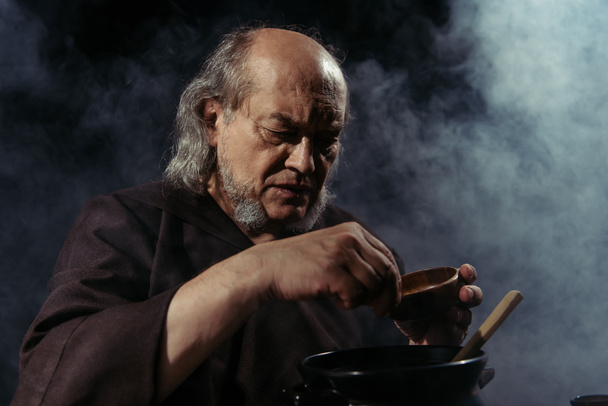 bearded alchemist adding ingredient into pot while preparing potion on black background with smoke - Photo, Image