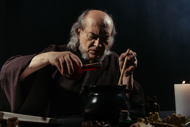 misterioso alquimista preparando poción mágica y vertiendo líquido en olla aislado en negro - Foto, Imagen