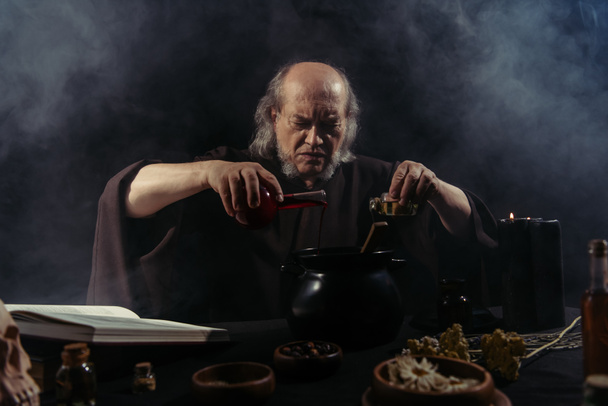 senior magician pouring liquid into pot while preparing potion on black background with smoke - Фото, зображення