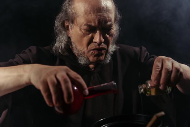 bearded alchemist mixing liquid ingredients and preparing magic potion isolated on black - Photo, Image