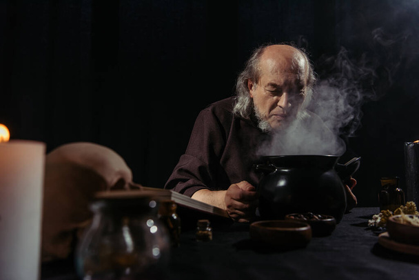 senior alchemist near boiling and steaming pot preparing potion at night isolated on black - Photo, Image