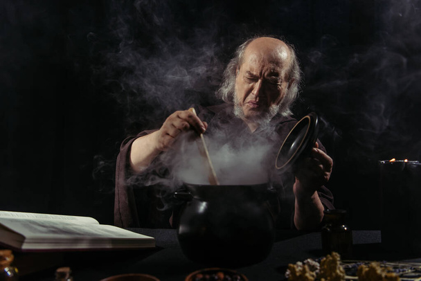 senior alkimista ráncolja homlokát közelében gőzölgő pot főzés közben éjjel fekete háttér - Fotó, kép