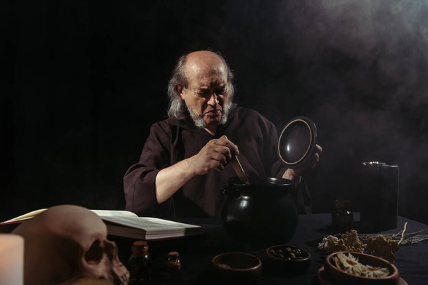 Mittelalterliches alchemistisches Stirnrunzeln in der Nähe dampfenden Topfes auf schwarzem Hintergrund  - Foto, Bild