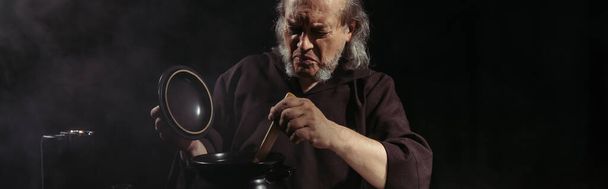 ανώτερος αλχημιστής ανάμειξη φίλτρο σε κατσαρόλα, ενώ το μαγείρεμα τη νύχτα απομονώνονται σε μαύρο, banner - Φωτογραφία, εικόνα