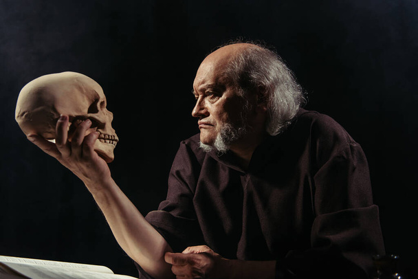 senior filosoof in donker gewaad kijkend naar menselijke schedel geïsoleerd op zwart - Foto, afbeelding