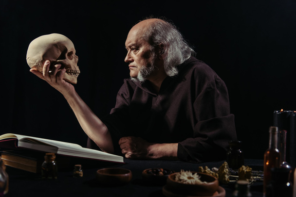 Senior-Alchemist schaut auf Schädel in der Nähe von magischen Kochbuch und pflanzlichen Zutaten isoliert auf schwarz - Foto, Bild