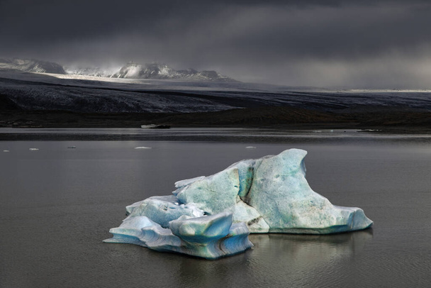 Fjallsarlon Glacier Lagoon, Iceland, on a stormy day. A blue iceberg floats in the lagoon and sunlight highlights the mountains behind. Part of the Vatnajokull National Park in Southern Iceland. - Fotoğraf, Görsel