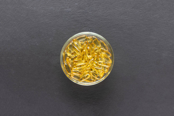 Cod liver fish oil capsules in glass bowl on black slate plate background. Omega 3, Vitamin E supplements. Healthy diet concept. Top view, copy space - Foto, imagen