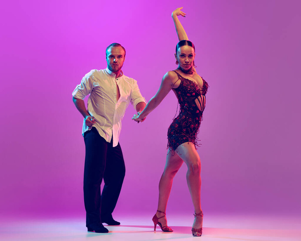 Elegance. Two young graceful dancers wearing stage outfits dancing ballroom dance isolated on purple background. Concept of art, dance, beauty, music, style. Copy space for ad - Φωτογραφία, εικόνα