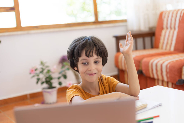 boy raises his hand during lesson Online distance learning. a boy with a computer communicate via videoconference with a teacher and a class group. The child communicates online. - 写真・画像
