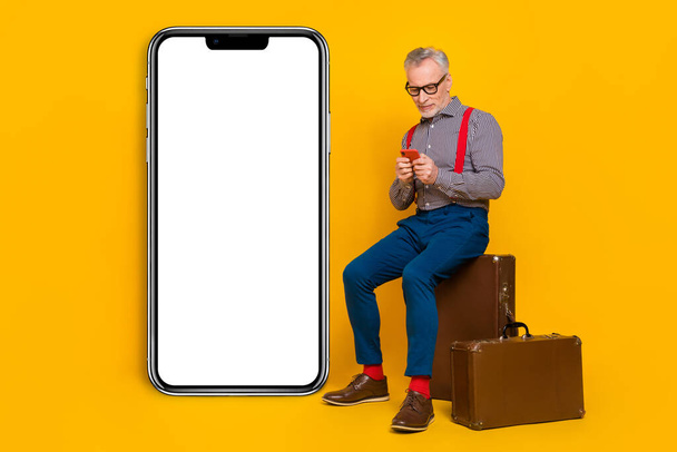 Full body photo of mature blogger waiting his flight in airport sitting baggage surfing smartphone isolated on yellow color background. - Fotoğraf, Görsel