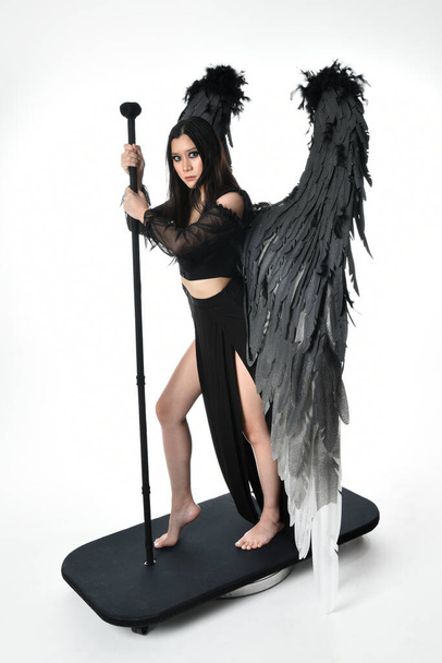 full length portrait of beautiful asian model with dark hair, wearing black gothic skirt costume, angel feather wings with horned headdress. Standing pose  isolated on studio background. - Foto, afbeelding