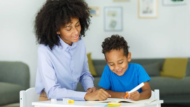 Female infant school teacher working one on one with a young schoolboy, sitting at a table writing in a classroom, front view. High quality photo - Foto, Imagen