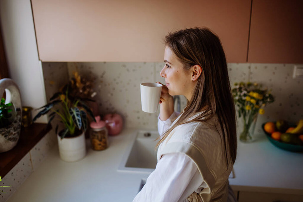 A smiling young woman with dreamy face drinking morning coffee at her kitchen. - Valokuva, kuva