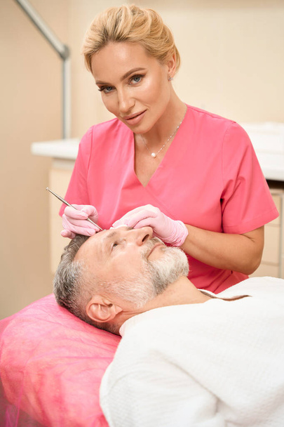 Smiling woman beautician with uno spoon cleans face of male patient in beauty salon. Cropped photo - Foto, imagen