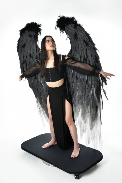 full length portrait of beautiful asian model with dark hair, wearing black gothic skirt costume, angel feather wings with horned headdress. Standing pose  isolated on studio background. - Foto, afbeelding