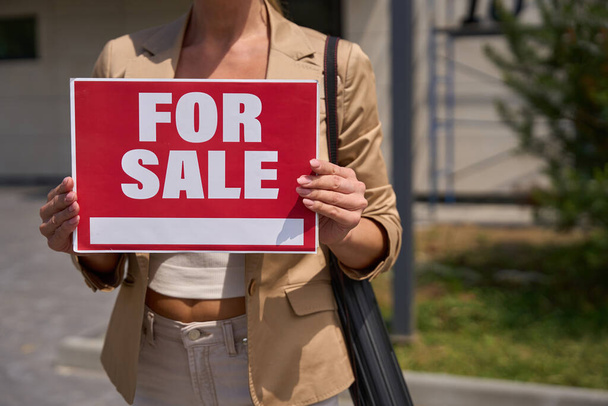 Female realtor stands with red sign in her hands for sale in front of house. Property for sale. Cropped photo - Foto, imagen