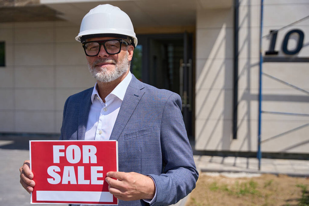 Focused foreman in a protective helmet holding a sign for sale. Man with glasses stands near a new house - 写真・画像