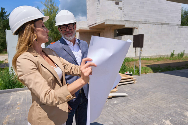 Real estate sales manager shows house project to buyer from the outside. Man and a woman in protective helmets - Valokuva, kuva