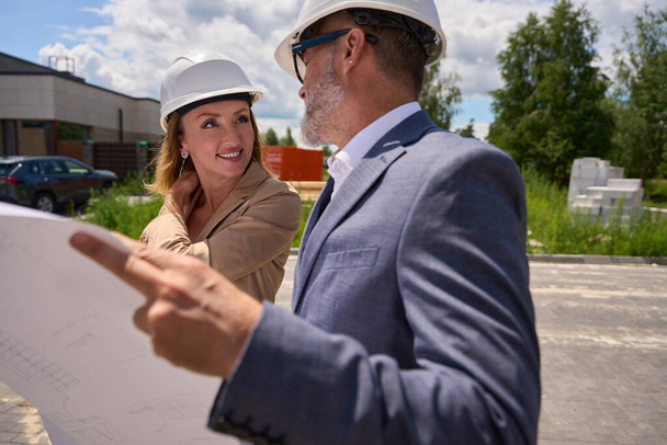 Woman realtor looks at client in a protective helmet who holds a drawing of house in her hands - Photo, image