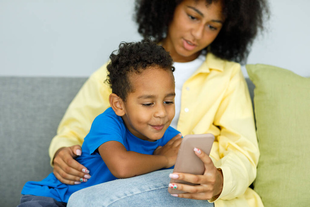 Little ethnic boy watching cartoons on phone smartphone with mother while sitting on couch at home together. High quality photo - Zdjęcie, obraz
