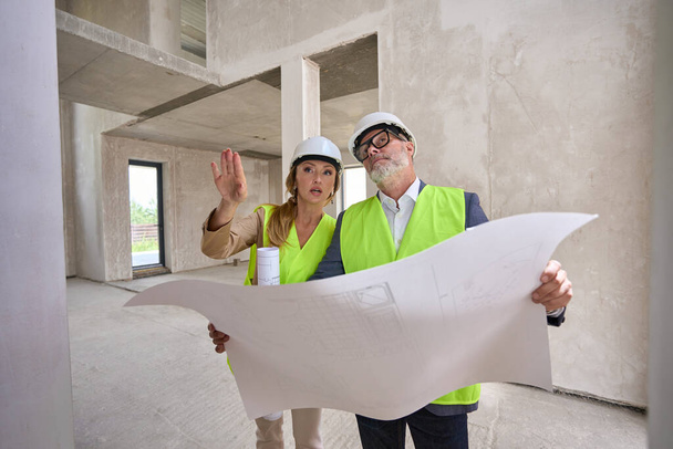 Foreman holds blueprint and discusses it with real estate manager. They stand inside an unfinished house - Fotoğraf, Görsel