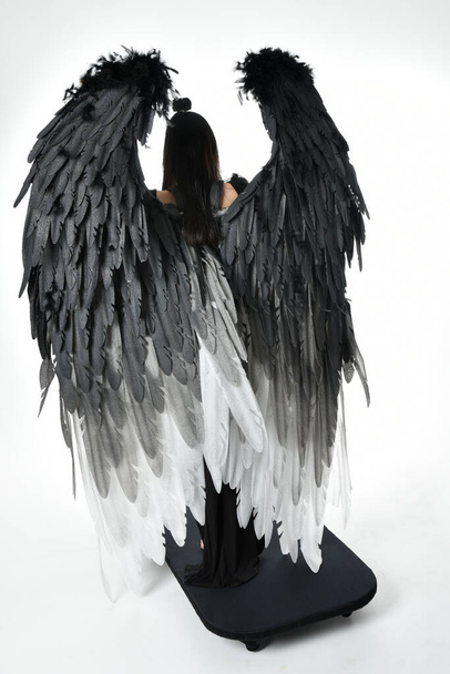 full length portrait of beautiful asian model with dark hair, wearing black gothic skirt costume, angel feather wings with horned headdress. Standing pose  isolated on studio background. - Fotó, kép