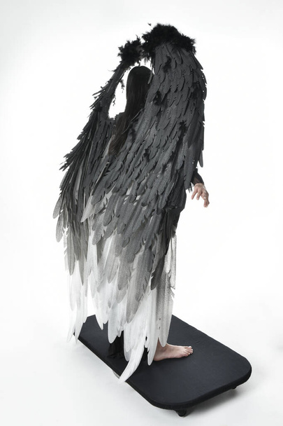 ολόσωμο πορτρέτο του όμορφου ασιατικού μοντέλου με σκούρα μαλλιά, φορώντας μαύρη γοτθική φούστα κοστούμι, φτερά αγγέλου φτερό με κέρατο κεφαλόδεσμο. Μόνιμη στάση απομονωμένη σε φόντο στούντιο. - Φωτογραφία, εικόνα