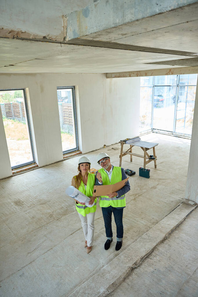 Realtor and foreman, who holds open folder in his hands, are discussing project of house. Couple stands inside unfinished building - Foto, Imagem