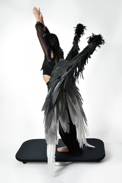 full length portrait of beautiful asian model with dark hair, wearing black gothic skirt costume, angel feather wings with horned headdress. Standing pose  isolated on studio background. - Photo, Image