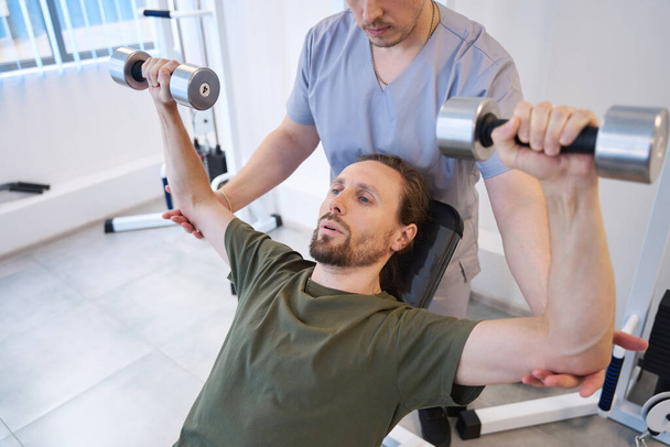 Patient diligently performs special exercises with dumbbells under the supervision of a doctor in a military sanatorium - Foto, Bild