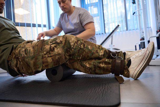Military patient does rehabilitation exercises under the supervision of a physiotherapist, he uses a special popliteal roller and fixation tourniquet - Photo, Image