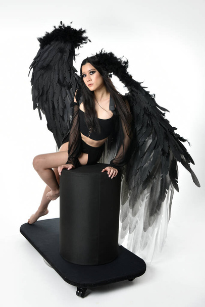 full length portrait of beautiful asian model with dark hair, wearing black gothic skirt costume, angel feather wings with horned headdress. Sitting pose  isolated on studio background. - Foto, imagen