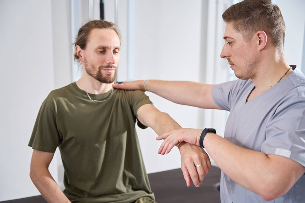 Military doctor in a military sanatorium helps an injured soldier develop a wounded arm in a session of physiotherapy exercises - Фото, изображение